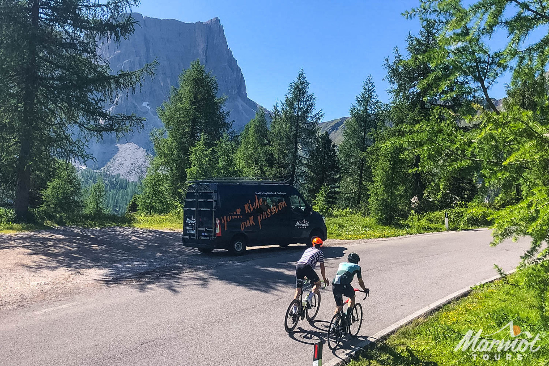 Pair of cyclist ride past Marmot Tours support vehicle on tree lines road with Dolomites backdrop on guided cycling holiday Italian Dolomites