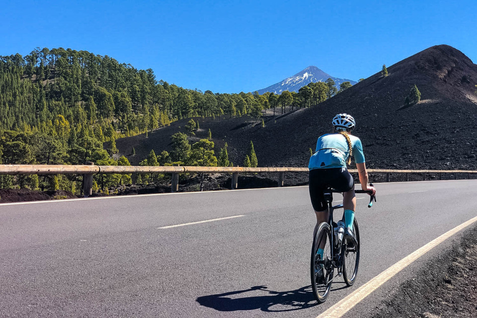Female cyclist climbing on smooth tarmac adjacent to lush pine forest with Mount Teide in background and a bright blue sky on Marmot Tours Tenerife cycling holiday